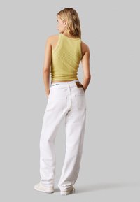 Top de tirantes amarillo con canalé y pantalones vaqueros de pierna ancha blancos. Los vaqueros tienen un parche de cuero marrón y dos bolsillos traseros. El calzado es de color claro.