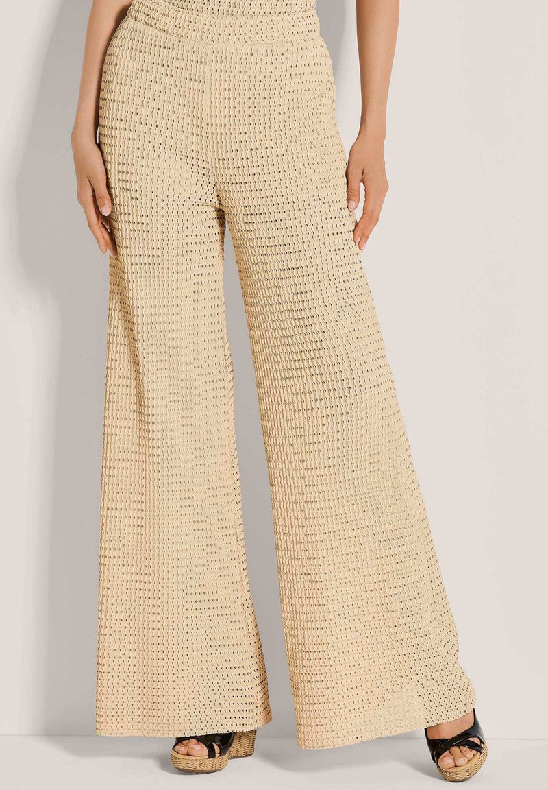 Beige weite Hose mit strukturiertem Strickmuster, getragen mit schwarzen Plateausandalen vor einem hellen Hintergrund.