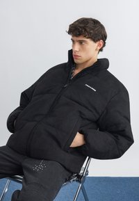 Junger Mann sitzt auf einem Metallstuhl, trägt eine schwarze Daunenjacke, die Hände in den Taschen, und schaut nach rechts vor einer schlichten weißen Wand.