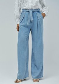 Pantalons en denim large à jambes droites de couleur bleu clair avec des plis, taille ceinturée, et poches latérales. Texture lisse avec une coupe décontractée, accompagnés de chaussures légères.