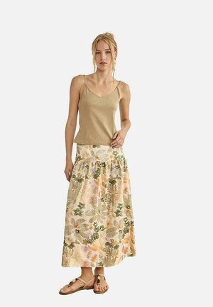Mujer con top sin mangas beige con tirantes de cadena, falda maxi con corte en A y estampado floral en tonos pastel, y sandalias planas marrones con cuentas decorativas.
