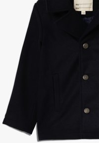 Námořnický vlněný peacoat s velkým výstřihovým límcem, třemi dekorativními knoflíky, boční kapsou a jemnou texturou. Podšívka uvnitř.