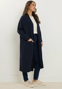 Lungo cardigan blu navy realizzato in morbido tessuto, con colletto a scialle, apertura frontale e due tasche anteriori. Indossato sopra una t-shirt bianca e jeans.