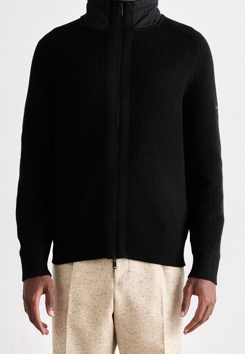 Pull noir côtelé zippé avec col montant porté sur un pantalon beige texturé.