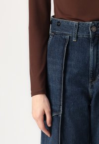 Ruskean pitkähihainen paita yhdistettynä sinisiin farkkulegginseihin. Legginsseissä on korkea vyötärö, nappiyksityiskohtia ja erottuvat tikkauskuviot.