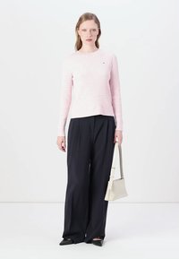 Pull en maille côtelée rose clair avec des manches longues, associé à un pantalon ample foncé et portant un sac à main blanc. Ballerines noires à bout pointu.