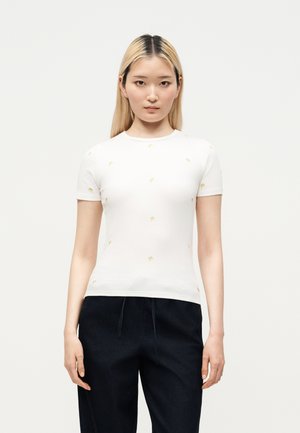 Jeune femme aux cheveux blonds et raides porte un haut blanc à manches courtes avec de petits accents jaunes et un pantalon foncé, debout devant un fond uni.