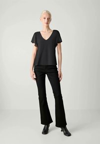 En blond kvinne står iført en svart V-hals bluse med gjennomsiktige korte ermer, svarte vide jeans og svarte ankelstøvler mot en grå bakgrunn.