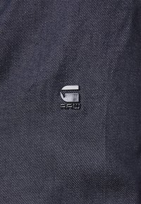 Dark blue fabric with a fine diagonal texture, featuring a shiny silver logo in the upper left corner. Minimalistic design, no patterns.