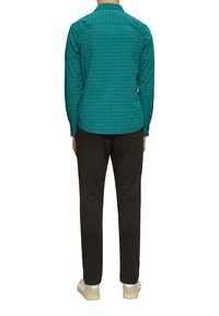 Turquoise lange mouwen shirt met een geometrische print, knoopsluiting bij de kraag en rechte zoom, gecombineerd met zwarte broek en lichtgekleurde schoenen.