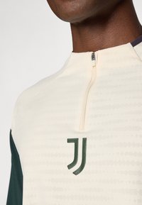 Cream zip-neck jersey with green accents. Features a textured pattern and a green logo on the left chest. Soft fabric, modern fit.