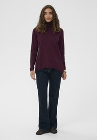Ženska s kodrastimi lasmi, oblečena v burgundski pulover s turtleneck izrezom, temno modre kavbojke in črne pletene balerinke, stoji pred enobarvno belo ozadje.