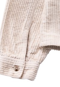 Beige corduroy jasje met verticale ribbels en een enkele doorzichtige knop op de mouw.