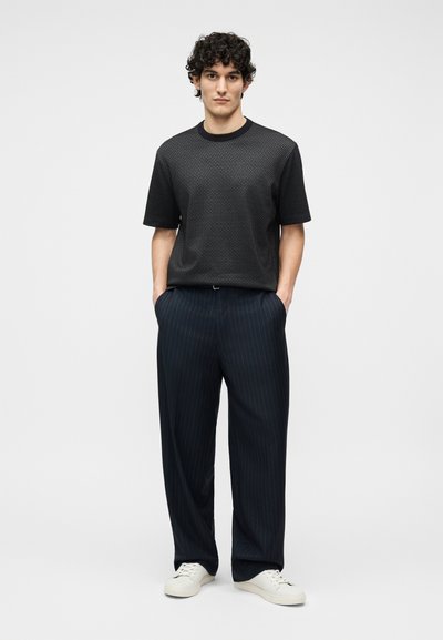 Νεαρός με κατσαρά μαλλιά, ντυμένος με μαύρο πουκάμισο κοντού μανικιού με σχέδια, μπλε παντελόνι με ρίγες και φαρδιά γραμμή, και λευκά αθλητικά παπούτσια, στέκεται με τα χέρια στις τσέπες.