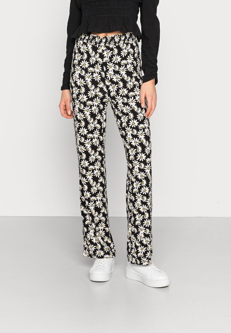Pantalons fleuris avec un fond noir, ornés de marguerites blanches à cœurs jaunes ; taille élastique, design à jambe droite, associés à des baskets blanches.