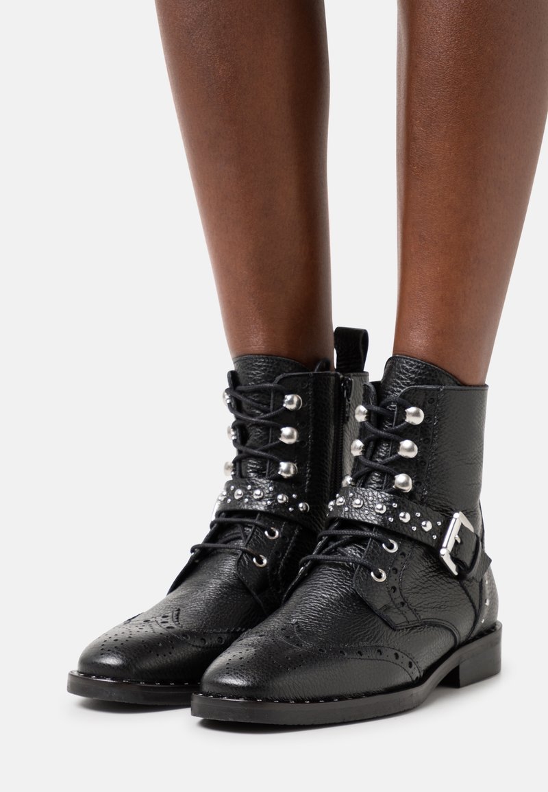 Bottes chevilles en cuir noir texturé avec détails brogue, clous argentés, sangle à boucle et lacets à l'avant portées sur des jambes à peau foncée.
