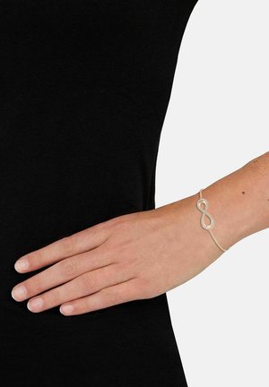 Pulsera de plata con un diseño de infinito, que presenta una cadena texturizada y una superficie lisa y pulida. Llevada en una muñeca desnuda sobre una tela negra.