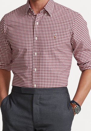 Homme portant une chemise à carreaux rouge et blanche à manches retroussées, un pantalon gris foncé et une montre-bracelet avec un bracelet en cuir marron.