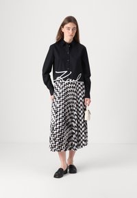 Camicia nera con bottoni e il logo bianco "Karl", abbinata a una gonna plissettata con un motivo a zigzag in bianco e nero. Scarpe slip-on nere.