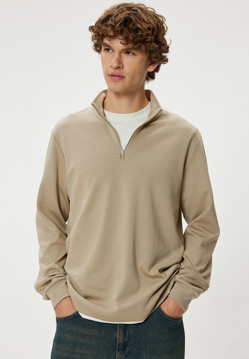 Pullover in pile beige con zip e maniche lunghe, trama morbida e vestibilità rilassata, indossato sopra una t-shirt bianca a girocollo.