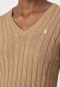 V-hals kabelgebreide trui in tan, met textuurpatronen. Een klein logo is zichtbaar aan de linkerkant. De stof is zacht en geribbeld.