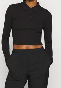 Schwarzes langärmliges verkürztes Poloshirt mit Kragen und einem Drei-Knopf-Verschluss, kombiniert mit hochgeschnittenen schwarzen Hosen. Strukturiertes Material.