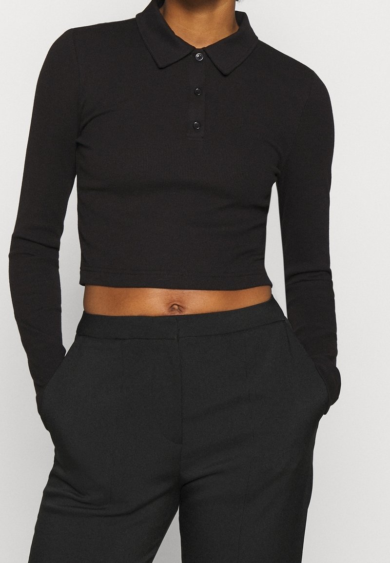 Schwarzes langärmliges verkürztes Poloshirt mit Kragen und einem Drei-Knopf-Verschluss, kombiniert mit hochgeschnittenen schwarzen Hosen. Strukturiertes Material.
