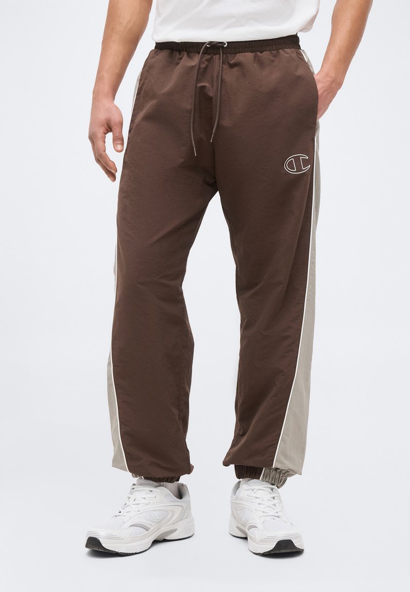 Bărbat purtând pantaloni de trening maro și bej cu talie cu șnur și adidași albi, stând cu mâinile relaxate pe lângă corp.