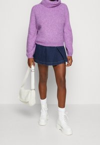 Personne portant un pull en maille violet, une jupe mini plissée bleu marine, des bottines blanches, des chaussettes blanches, et tenant un sac à main en maille blanche.