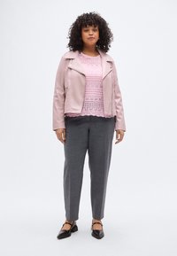 Hellrosa Jacke aus Kunstleder über einem kurzärmligen Top mit Spitzenmuster; graue, maßgeschneiderte Hose; schwarze Schuhe mit Knöchelriemen.