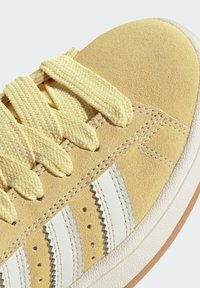 Zapatillas de ante amarillas con rayas blancas, cordones texturizados y suela de goma. Destacan los detalles de costura a lo largo de los bordes.