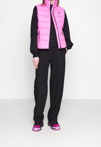 Person, die eine leuchtend pinke Steppweste über einem schwarzen Langarmshirt, schwarzen Hosen und schwarz-pinken Sneakers trägt, steht mit den Händen in den Taschen.