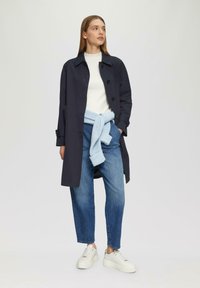 Marineblauwe trenchcoat met knopen, gedragen over een witte geribde trui. Lichtblauwe gebreide trui geknoopt om de taille, gecombineerd met spijkerbroek en witte sneakers.