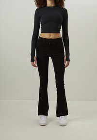 Personne debout portant un haut court noir à manches longues, un pantalon noir évasé et des baskets blanches sur un fond uni.