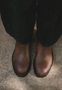 Bottes basses en cuir marron à finition lisse, bout arrondi et détails de couture contrastants, portées avec un jean foncé.