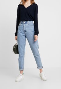 Granatowa bluzka z długim rękawem i dekoltem w serek połączona z jasnoniebieskimi spodniami typu high-waisted oraz białymi trampkami, trzymająca małą, okrągłą czarną torebkę.