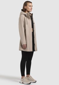 Veste imperméable beige avec capuche, accents noirs, zippers et cordons de serrage. Associée à des leggings noirs et des baskets beiges.