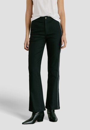 Moteris dėvi juodas prigludusias kelnes su baltomis šoninėmis juostelėmis, baltą rankovių neturintį viršų ir juodus aukštakulnius aulo batus su smailiu noseliu, stovinčia pilkoje fone.