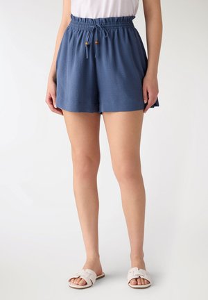 Kobieta nosząca niebieskie krótkie spodenki z elastycznym pasem i sznurkiem oraz białe sandały wsuwane, stojąca na tle jednolitego jasnego tła.