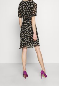 Robe noire à imprimé floral avec des manches courtes, taille cintrée et jupe fluide. Portée avec des escarpins en daim violet. Texture lisse.