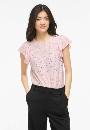 Femme aux longs cheveux foncés portant un haut rose clair à manches papillon ajourées et un pantalon noir, debout les mains dans les poches sur fond blanc.