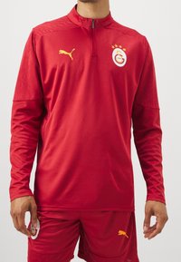 Rød langærmet sportstrøje lavet af glat stof, med høj krave, lion logo og gule accenter. Vist med matchende shorts.