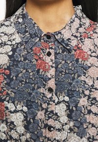 Camicia a bottoni floreale con sfondo blu scuro adornato da fiori rosa, rossi e blu. I bottoni neri aggiungono contrasto al design.