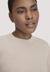 Beige gerippter Strickpullover mit Rundhalsausschnitt, der ein strukturiertes Muster und einen subtilen Glanz aufweist. Nahaufnahme des Stoffes und des Ausschnitts.