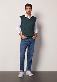 Zielony, dzianinowy kamizelka noszona na białej koszuli z kołnierzykiem, w połączeniu z niebieskimi dżinsami i białymi trampkami. Design z dekoltem w serek oraz bez rękawów.