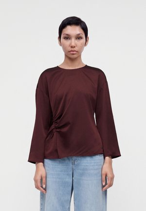 Femme aux cheveux courts et foncés portant un haut à manches longues marron foncé avec fronces latérales et un jean bleu clair, debout devant un fond blanc uni.