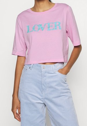 Osoba nosząca różowy krótki T-shirt z napisem "LOVER" niebieskimi literami oraz jasnoniebieskie, sztruksowe spodnie z wysokim stanem, jedna ręka w kieszeni.