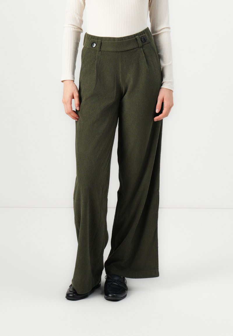 Pantalons en velours côtelé vert olive à jambes larges avec une texture côtelée, dotés de deux boutons à la taille et de poches latérales. Associés à des chaussures noires.