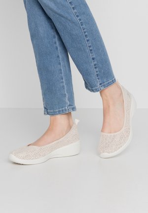 Füße tragen beigen Slip-On-Keilabsatzschuhe mit Spitzenmuster, kombiniert mit blauen Jeans vor einem schlichten hellen Hintergrund.