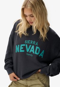 Sudadera gris oscuro con "SIERRA NEVADA" en letras turquesa. Presenta un corte holgado, mangas largas y una textura de tela suave.
